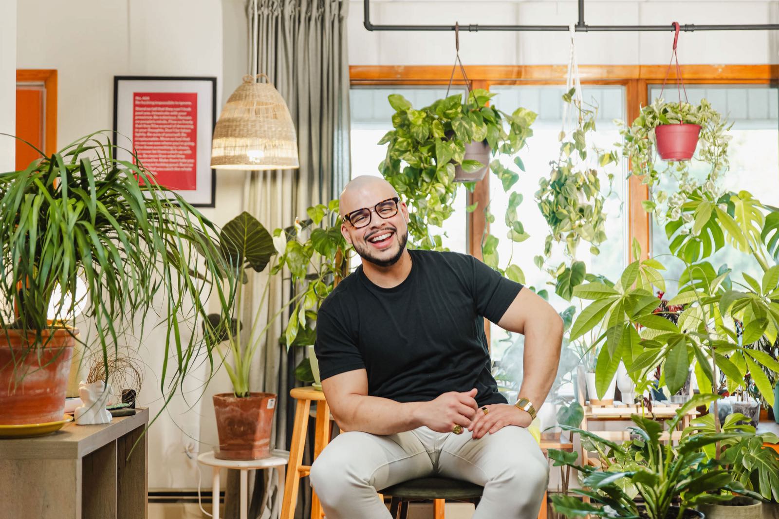 Willis Aybar, founder of Another Monday, laughing on a stool in his plant-filled room at home in Baltimore, MD, where he works out the local marketing strategies this guide walks through