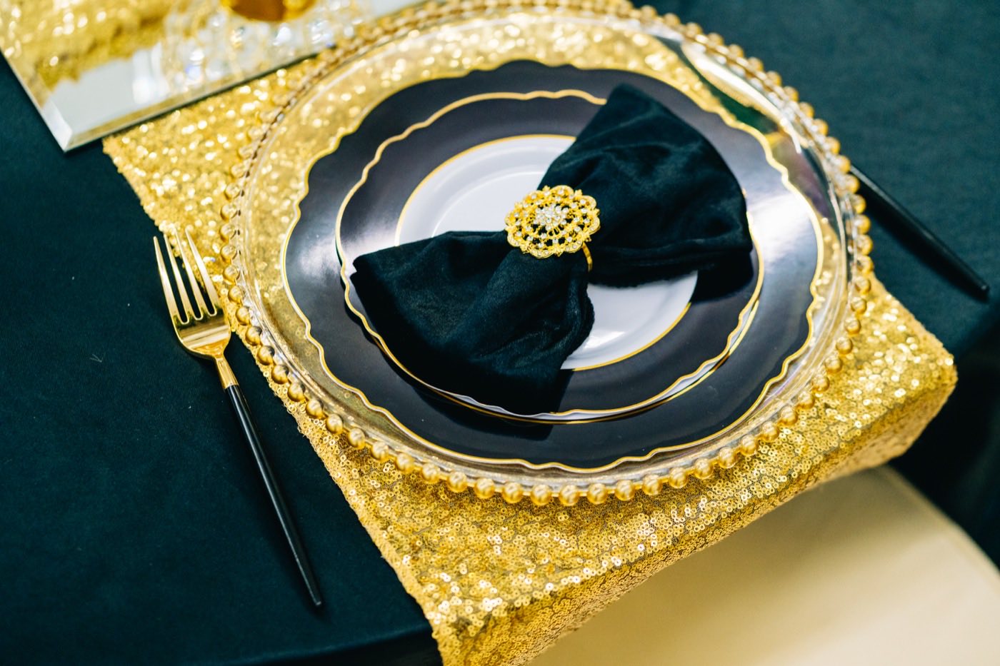A black and gold place setting styled with a velvet napkin and gold flatware on a sequin table runner, the kind of styling detail a venue's premium tier covers