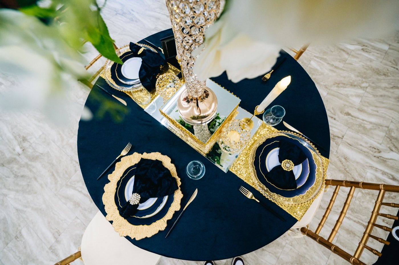 Overhead view of a round table styled at Studio 87 with navy linen, gold beaded chargers, black napkins, a crystal candelabra centerpiece and gold chiavari chairs, a table set and ready before guests arrive