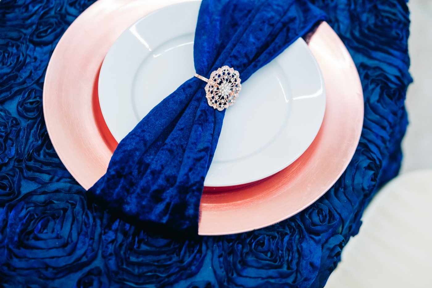 An overhead place setting styled with a navy velvet napkin, a rose-gold charger and a rhinestone napkin ring on a navy rosette linen, the kind of real styling photo couples shortlist a venue on
