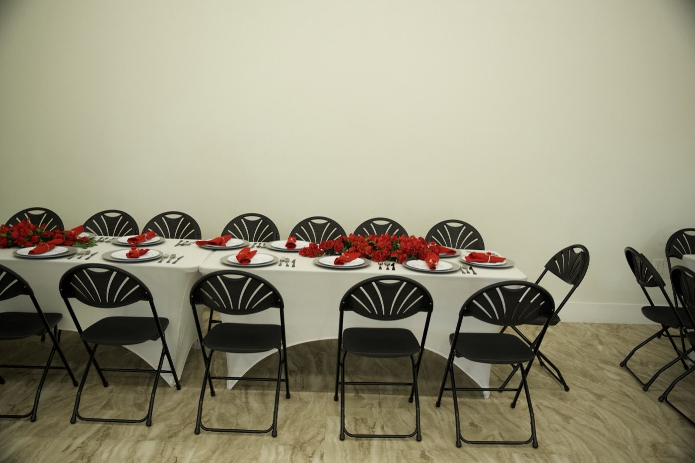 A long banquet table at Studio 87 set with white plates, red rose centerpieces and red napkins, flanked by black fan-back folding chairs against a plain white wall, the kind of styled photography a venue site needs before anyone books a tour