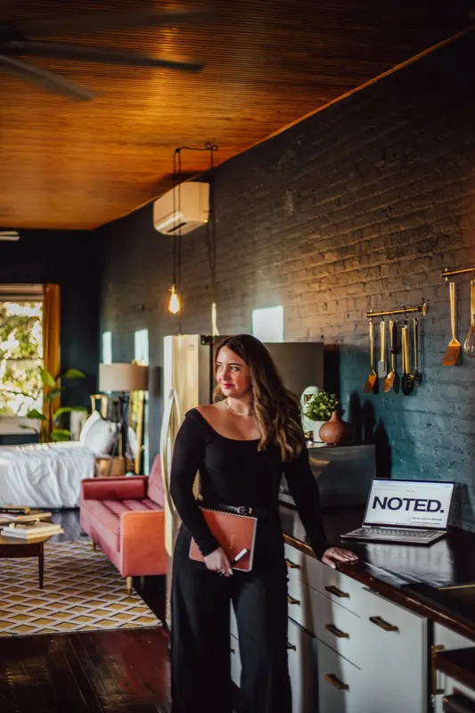 Allison Chavis of Noted., a Baltimore virtual assistant, standing in a loft kitchen with a planner beside a laptop showing the Noted. logo